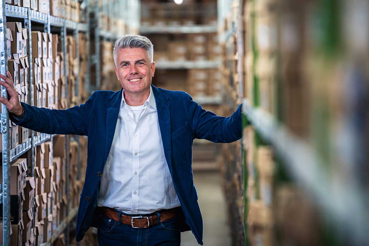 Confident businessman in a blue blazer and white shirt standing in a warehouse aisle between shelves filled with boxes, smiling at the camera