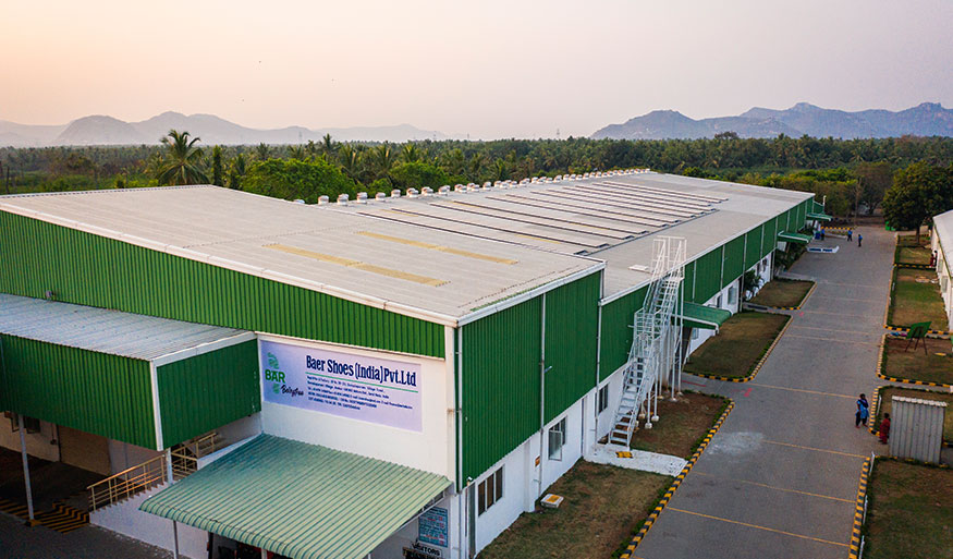 Aerial shot of an industrial building with a green roof, located at Baer Shoes India Pvt Ltd, surrounded by a road and greenery, with mountains in the background.