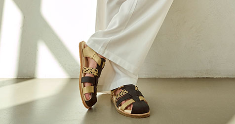 Person wearing stylish leather sandals with brown and beige straps and white pants, standing indoors