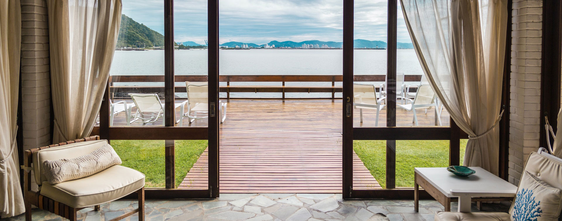 Living room with a view of the ocean through large glass doors, leading to a wooden patio with chairs.
