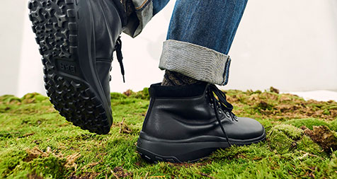 A person wears black ankle boots with rugged soles, standing on a mossy surface, showcasing outdoor style and comfort.