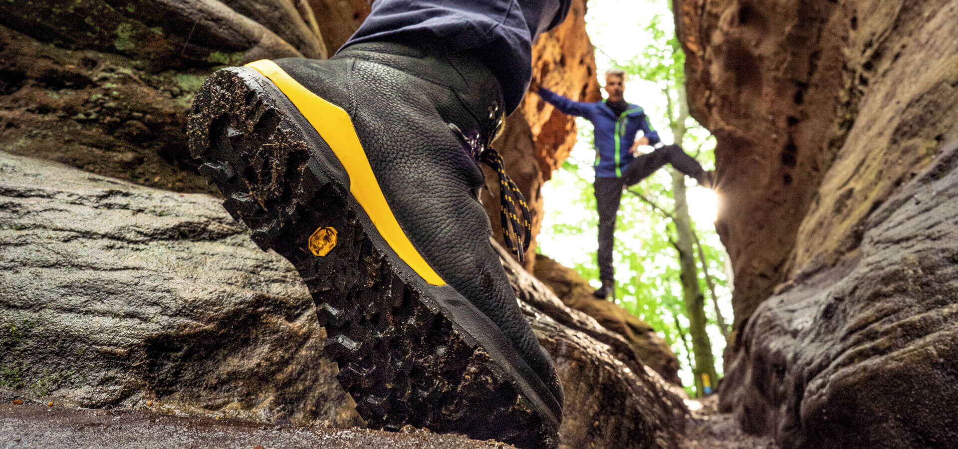 Close-up of a rugged hiking boot with yellow accents on a muddy, rocky trail, with a person climbing in the background.