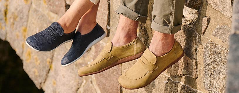 Close-up of two pairs of shoes, navy slip-on sneakers for women and tan loafers for men, worn by people sitting on a stone wall
