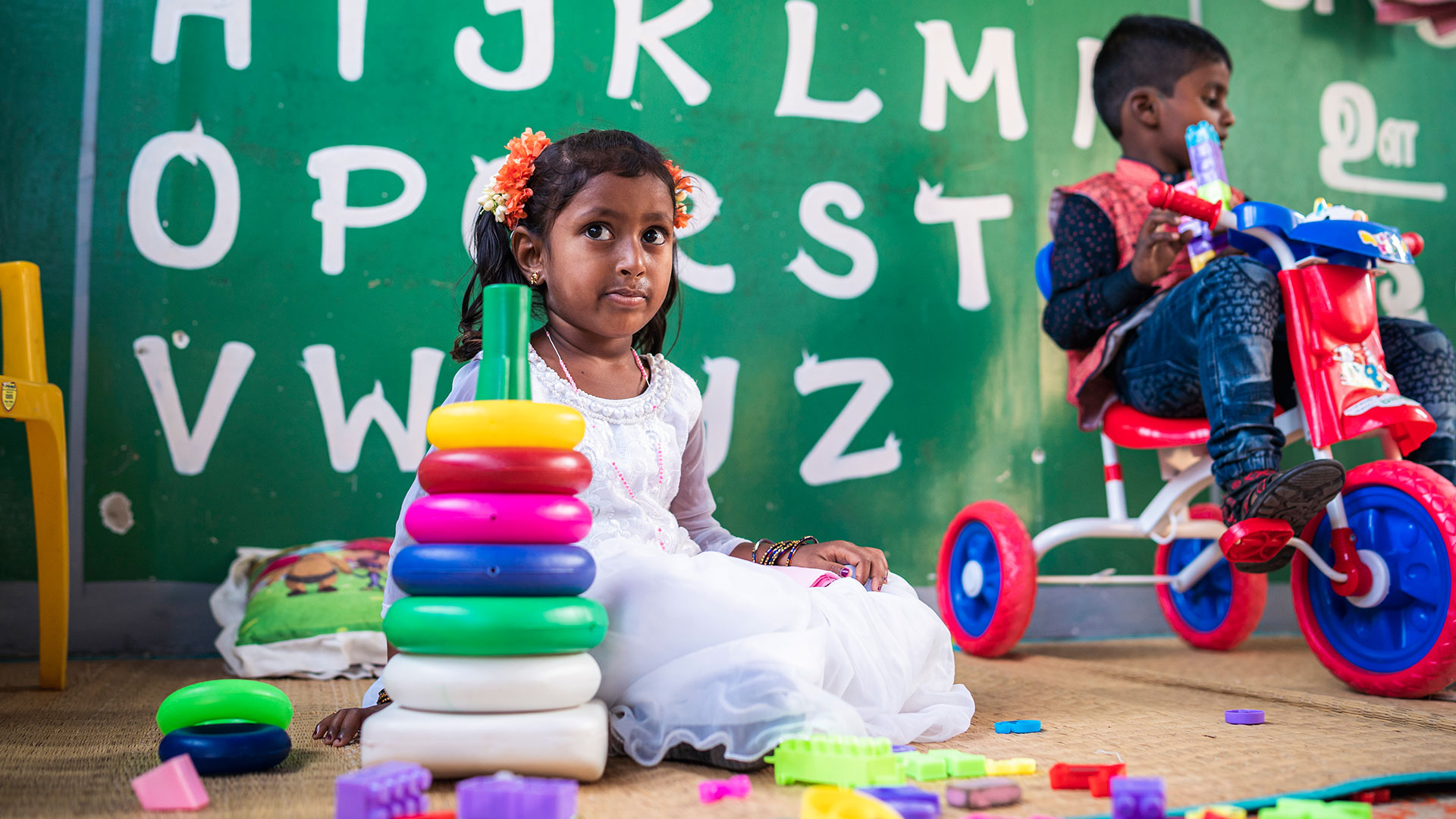 Two children are in a classroom with alphabet letters on the wall. A girl in a white dress plays with colorful stacking rings, while a boy on a red tricycle holds a toy. The scene is lively with various educational toys around.