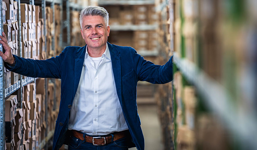 Smiling man in a blue jacket and white shirt in a warehouse setting, surrounded by shelves and boxes
