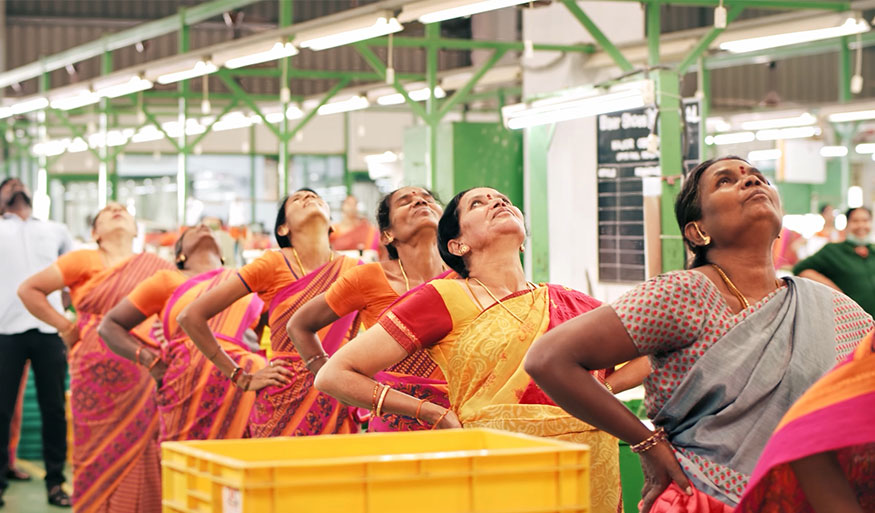 A group of women in vibrant sarees doing stretching exercises together in a factory environment with a focus on health and well-being at work