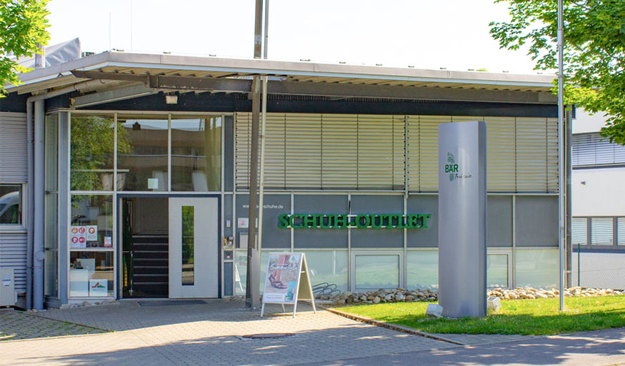 Front view of a modern shoe outlet with large windows and green signage, surrounded by trees on a sunny day