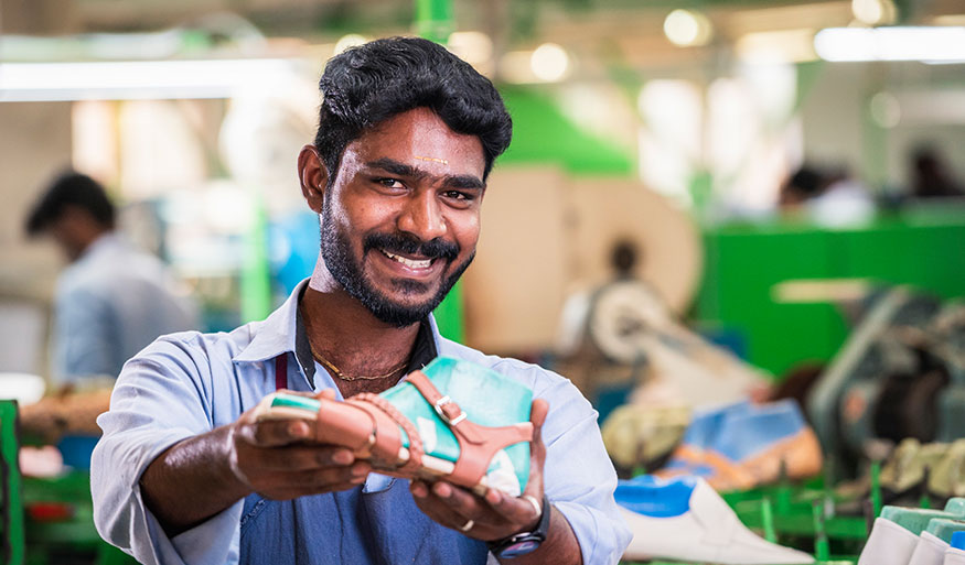 A smiling man in a factory setting presents a shoe, showcasing craftsmanship and quality in footwear production.