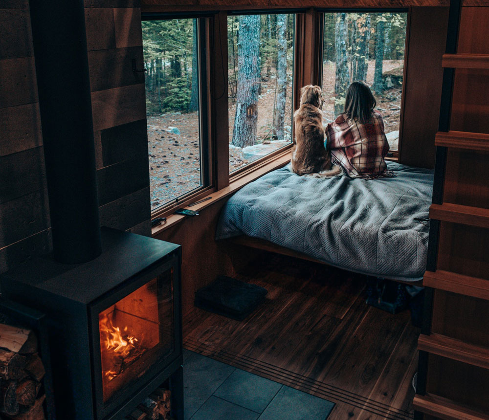 A woman and her dog sitting on a bed in a wooden cabin, looking out the window at a forest view, next to a warm fireplace.
