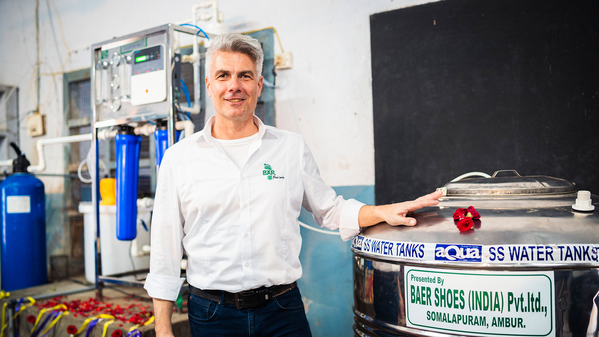 Man in white shirt standing beside a large stainless steel water tank with filtration system in background