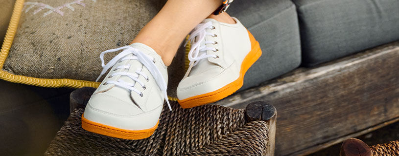 Person wearing stylish white sneakers with orange soles on a woven seat