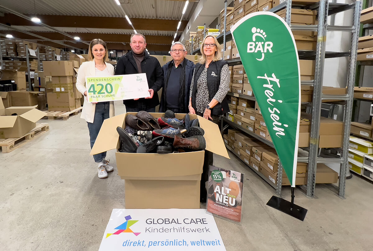 Group of people with a donation box of shoes for charity by BÄR Footwear, displaying a sign reading 'Donation Certificate 420 Pairs of Shoes', set in a warehouse.