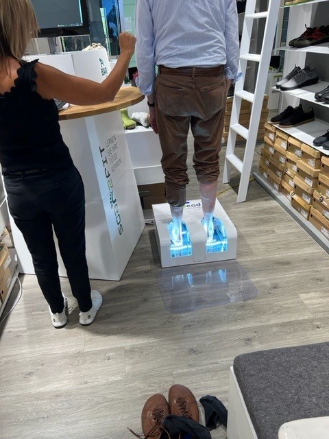 A man standing on an advanced foot measuring scanner in a shoe store, assisted by an employee.