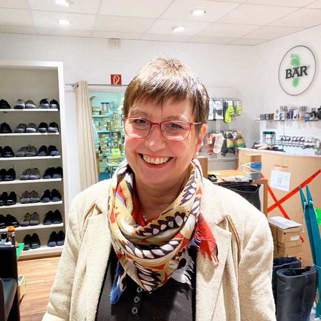 A woman with short hair and glasses smiling in a shoe store, wearing a colorful scarf and surrounded by shelves and boxes.
