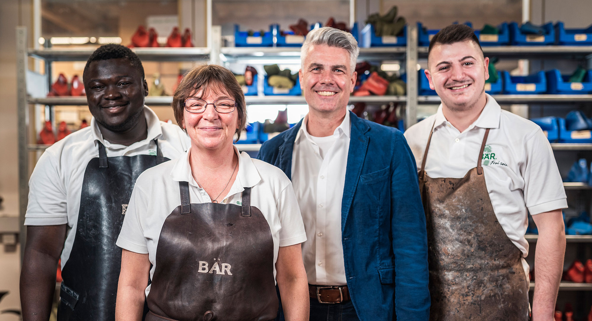 Group of four people standing in a workshop with shoe molds on shelves, all wearing aprons and smiling at the camera