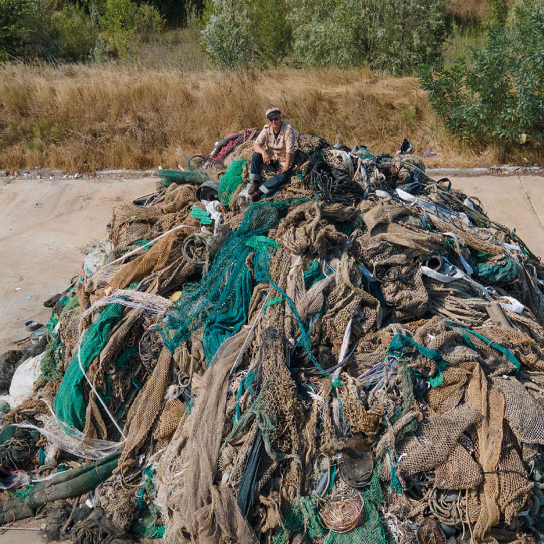 A person is sitting on top of a huge pile of old, tangled fishing nets in an open area with grassy surroundings.
