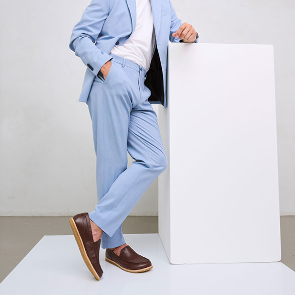 Man wearing a stylish light blue suit and brown slip-on shoes, leaning casually against a white pedestal