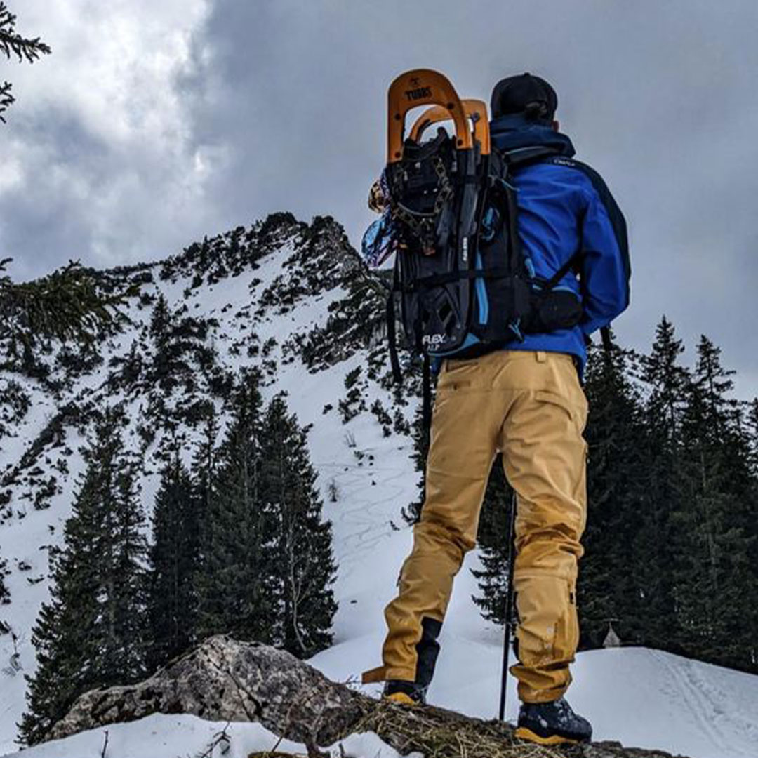Person in outdoor gear stands on snowy mountain with snowshoes on backpack, surrounded by trees and rugged peaks